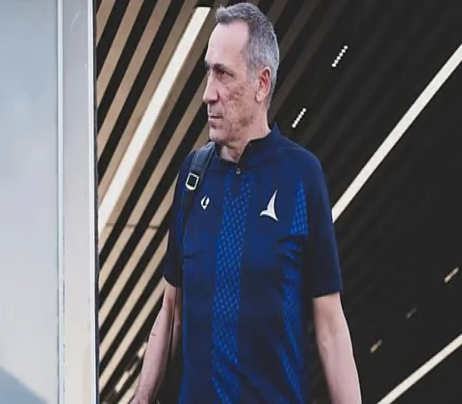 رجل يرتدي قميصاً رياضياً أزرق ويحمل حقيبة على كتفه، يسير في ممر ذو خلفية مخططة.